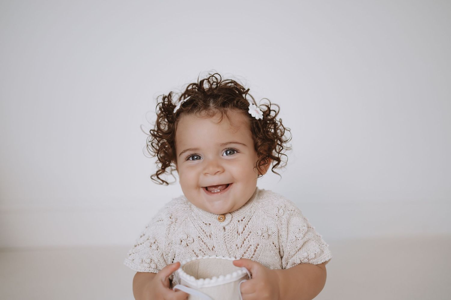 A baby wearing a white lace dress sits on a white backdrop while smiling and laughing during a photo session.