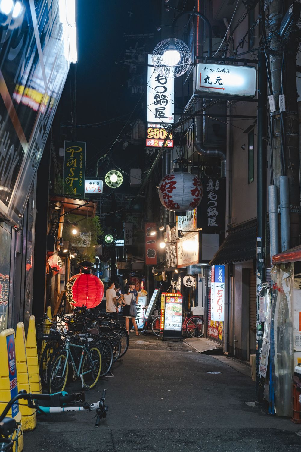 Dimly lit back alley in Japan with red lanterns, bicycles, and vending machines.