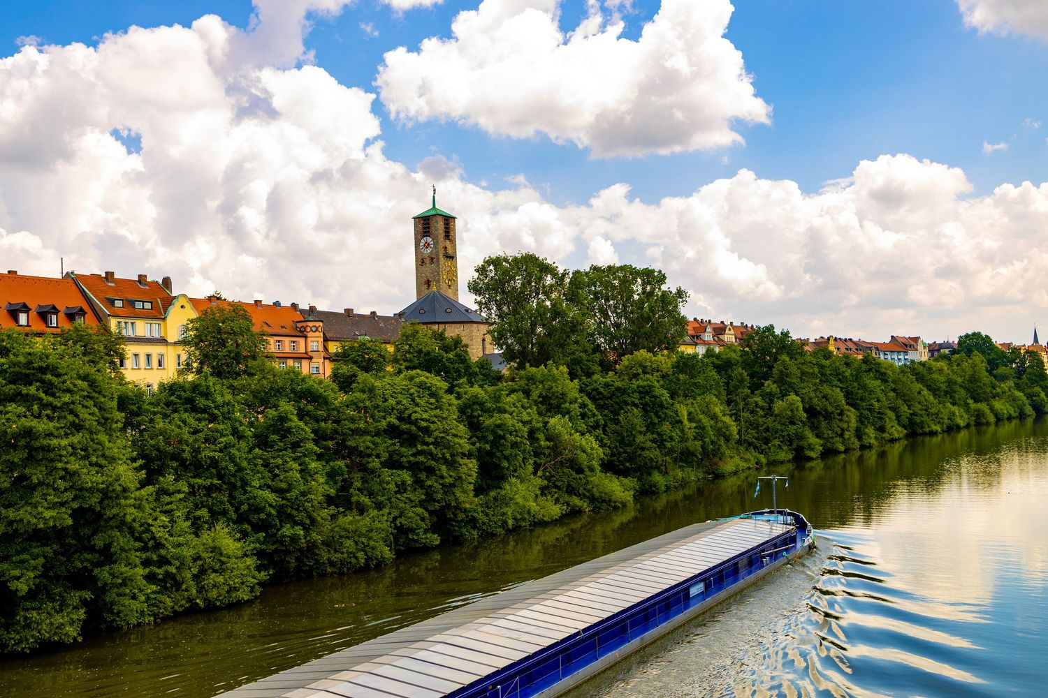 Scenic waterway with historic buildings and boats along tree-lined riverbank.