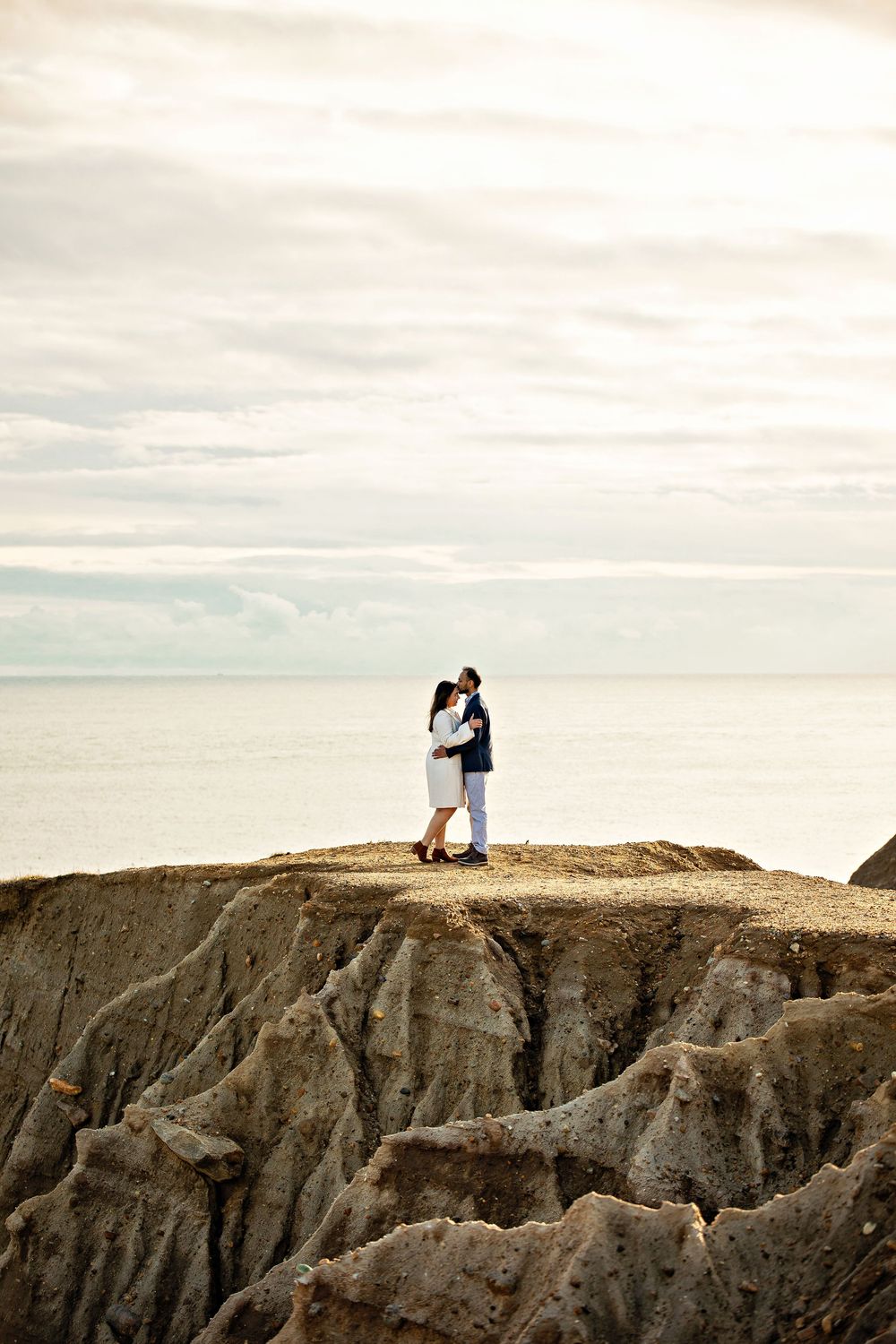 Two people stand on a dramatic rocky cliff edge overlooking a vast landscape under cloudy skies.