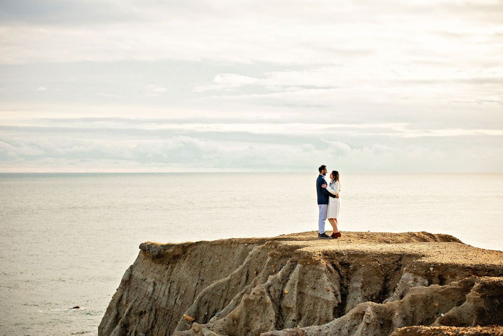 Two silhouettes stand atop dramatic coastal cliffs overlooking a misty ocean at sunset.