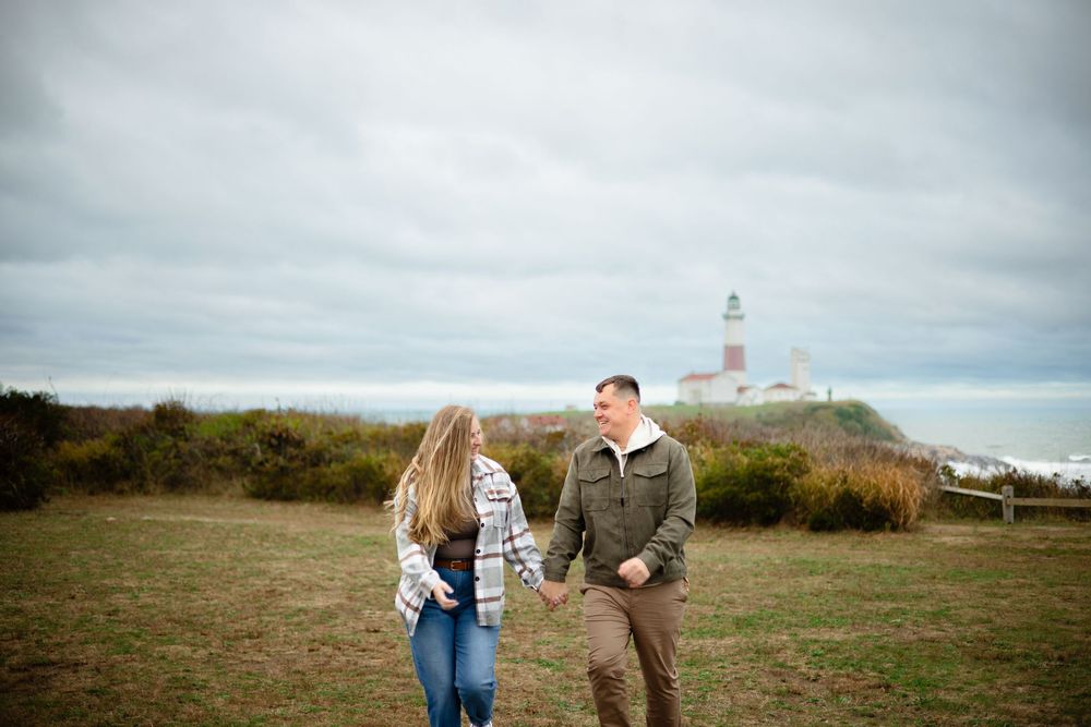 A couple walks hand in hand through a scenic coastal landscape with a lighthouse in the distance.