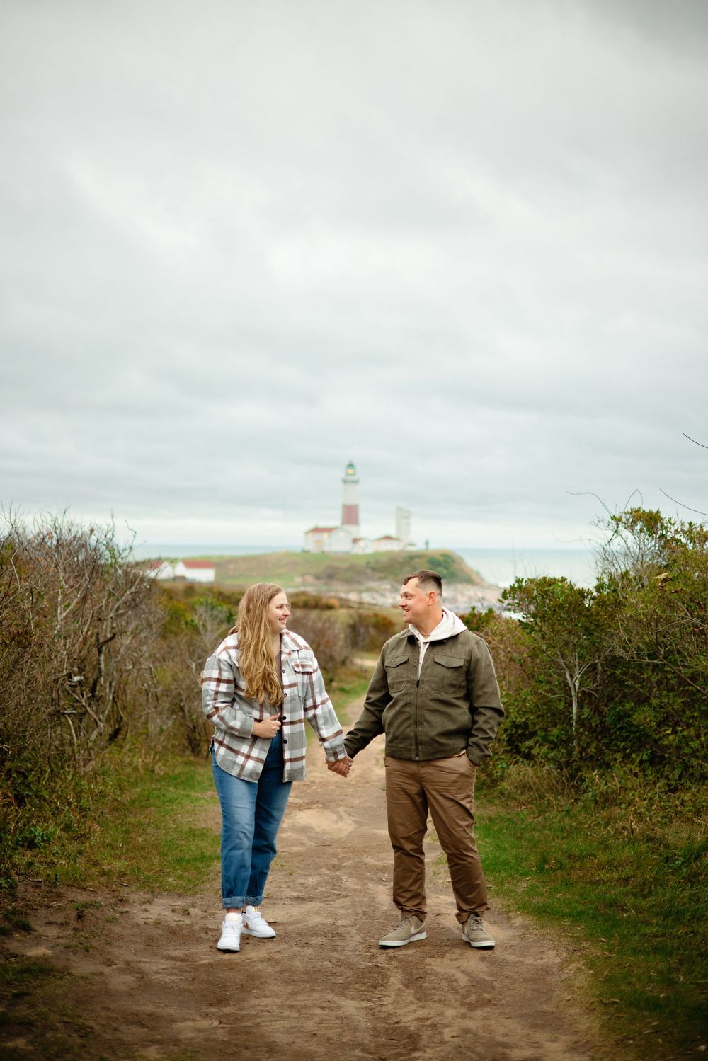 Two people walking and laughing together on a coastal trail with a lighthouse visible in the distance on an overcast day.