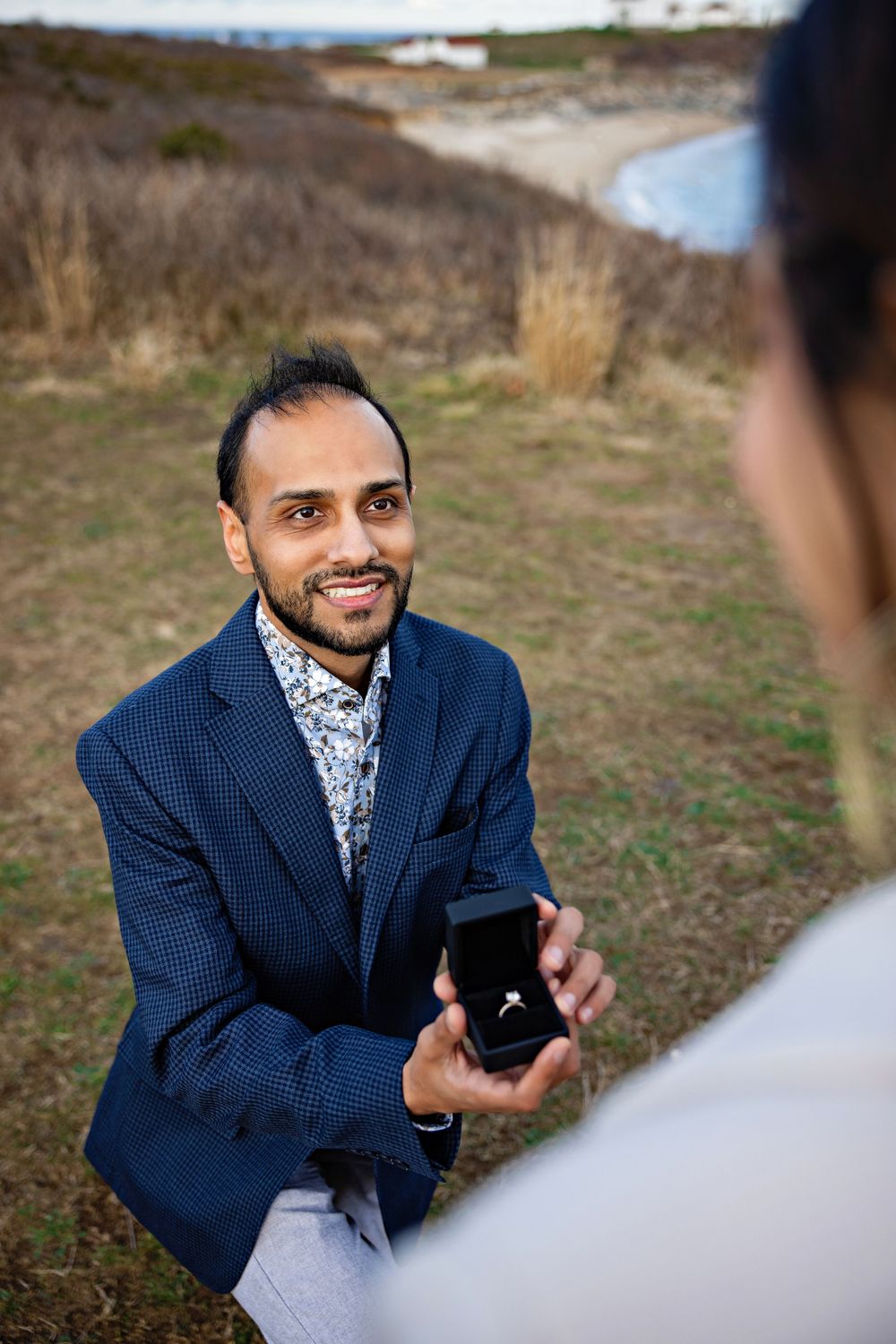 A person in a navy suit jacket and patterned shirt kneels while holding a small box outdoors.