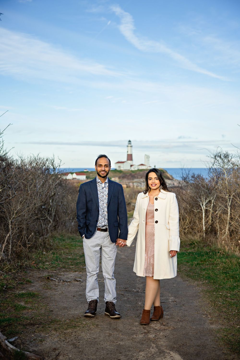 A lighthouse stands on the horizon as a couple walks along a coastal path on a clear winter day.