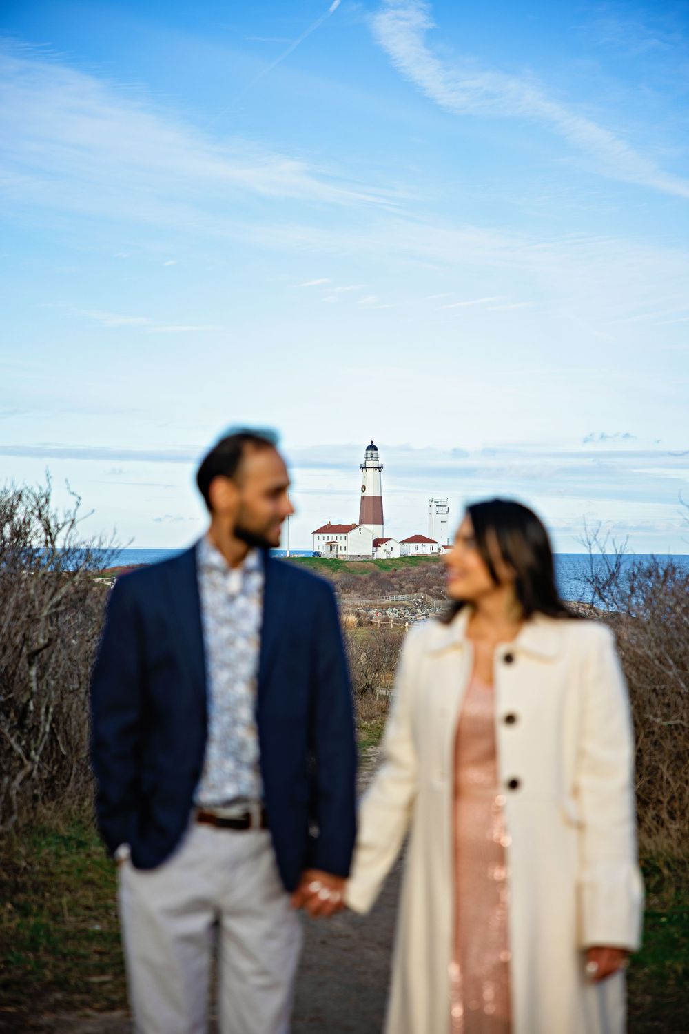 A romantic couple poses in front of a lighthouse on a bright sunny day with blue skies and coastal scenery.