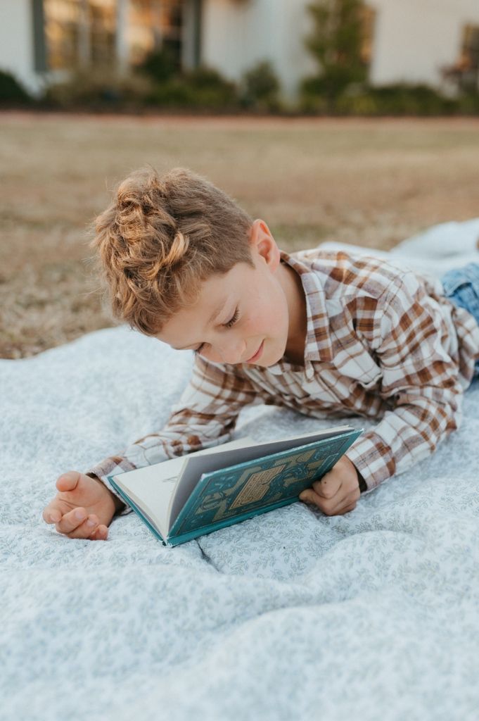 Child resting on white blanket while reading a book outdoors.