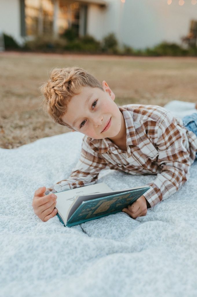 A child in a plaid shirt reading a book while lying on a white blanket outdoors at sunset.
