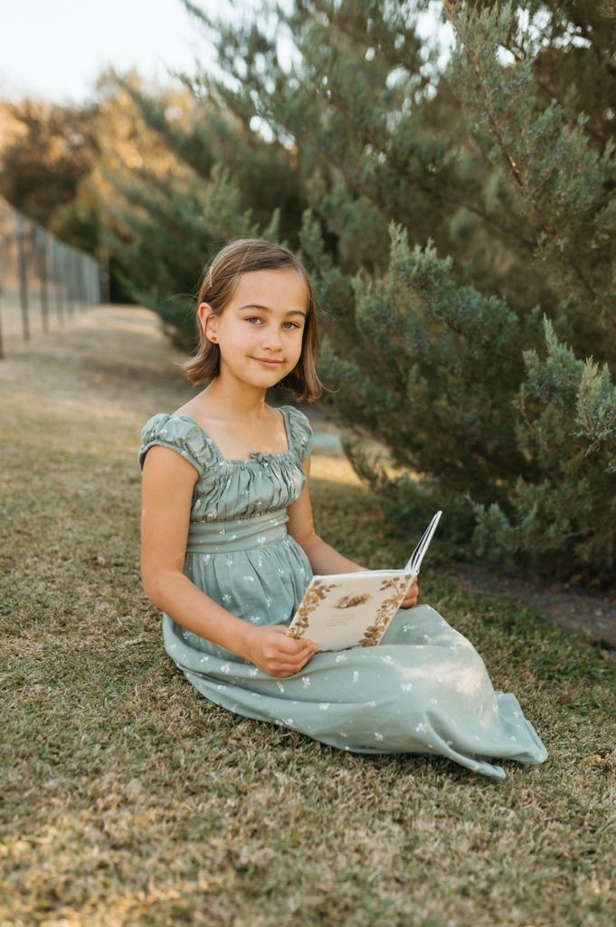 Young reader in teal dress enjoys a book while sitting on grass near evergreen bushes.