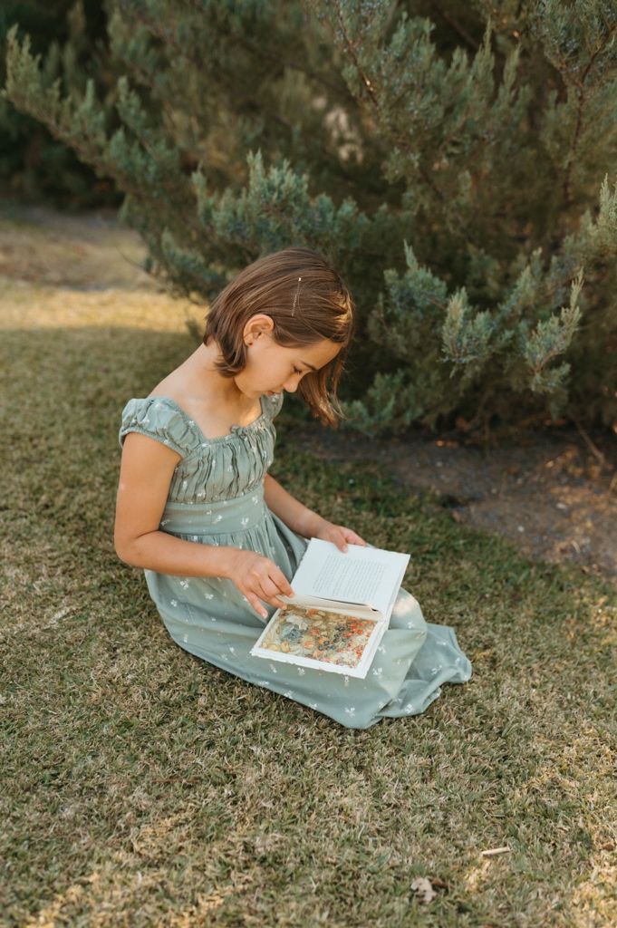 Young child in a blue dress sits on grass reading a picture book outdoors.