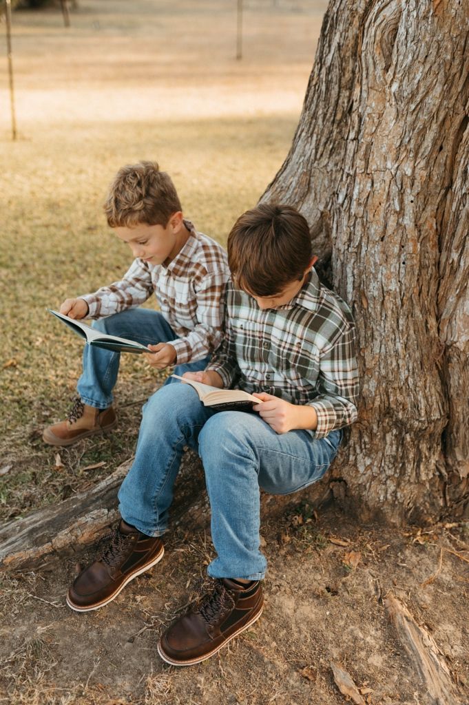 Young child sits against tree trunk while reading or writing in notebook on autumn day.