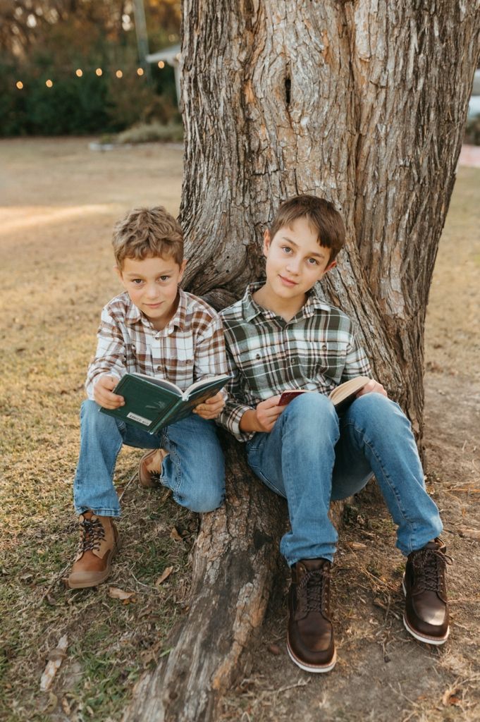 Two children reading books while sitting against a large tree trunk in an outdoor setting at sunset.