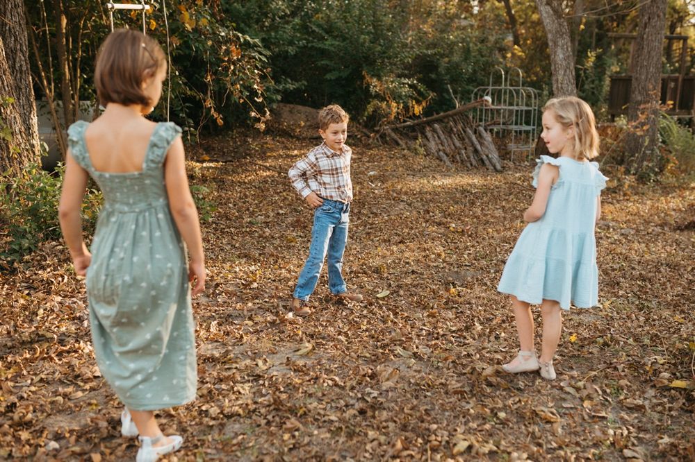 Children in blue dresses play and explore together in a sunlit autumn forest setting with fallen leaves.