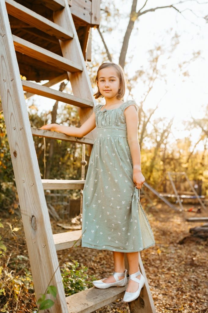 Child in mint green floral dress stands on wooden steps in golden sunset light among autumn leaves.