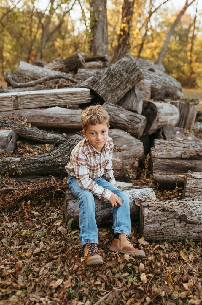 A child in a plaid shirt and jeans sits on a woodpile surrounded by autumn leaves and trees in golden light.