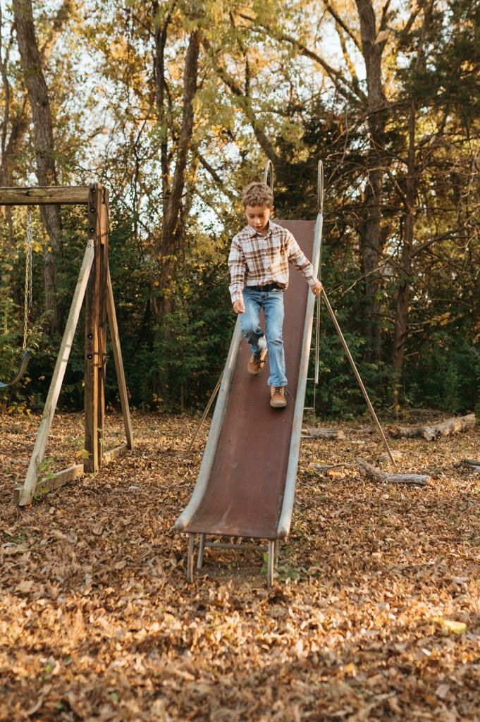 Child plays on a wooden slide surrounded by autumn leaves in a wooded outdoor setting.