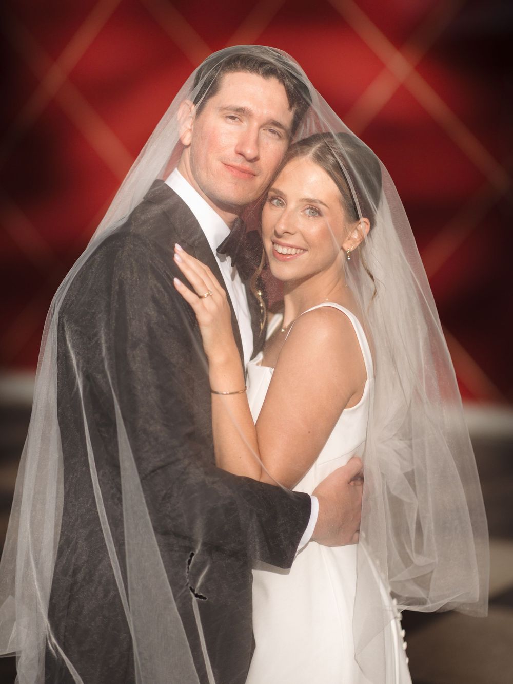 A bride and groom pose together for a formal wedding portrait against a red backdrop with soft lighting.
