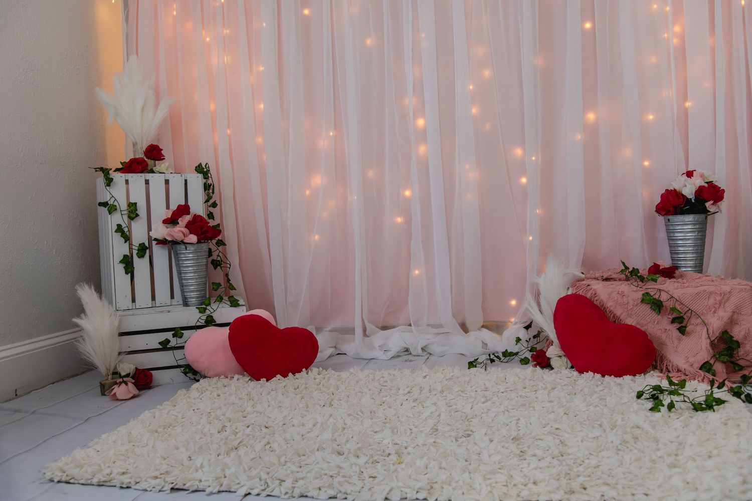 Romantic Valentine's Day photo backdrop with white curtain lights, red hearts, roses, and plush white carpet.