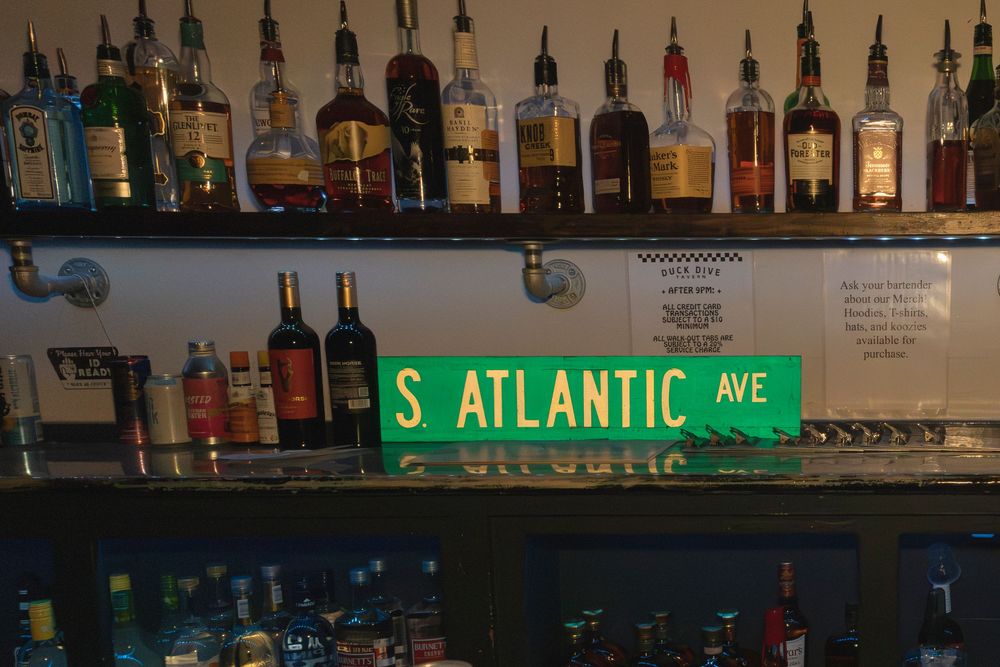 Bar shelf stocked with liquor bottles under ambient lighting with street sign visible.