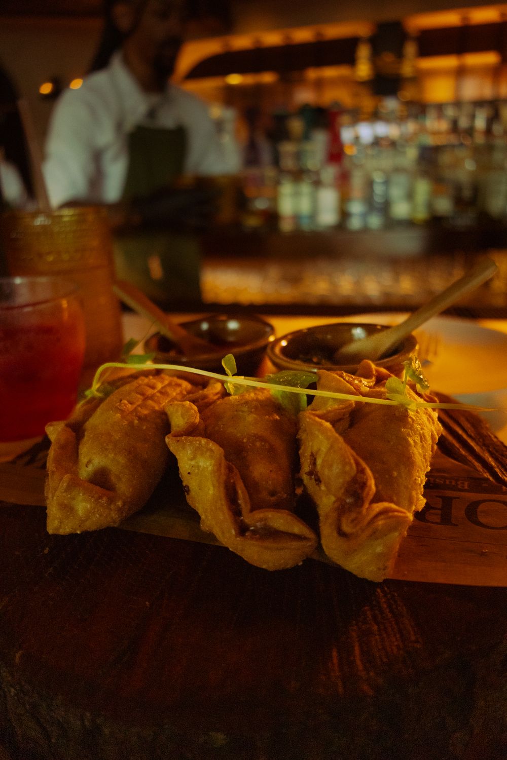 Row of gourmet tacos arranged on a wooden serving board in warm restaurant lighting.