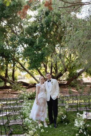A couple poses in formal attire under blooming trees in a garden setting.