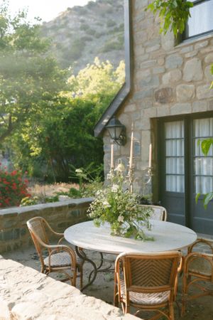 Rustic outdoor patio setting with white round table and wicker chairs against stone wall.