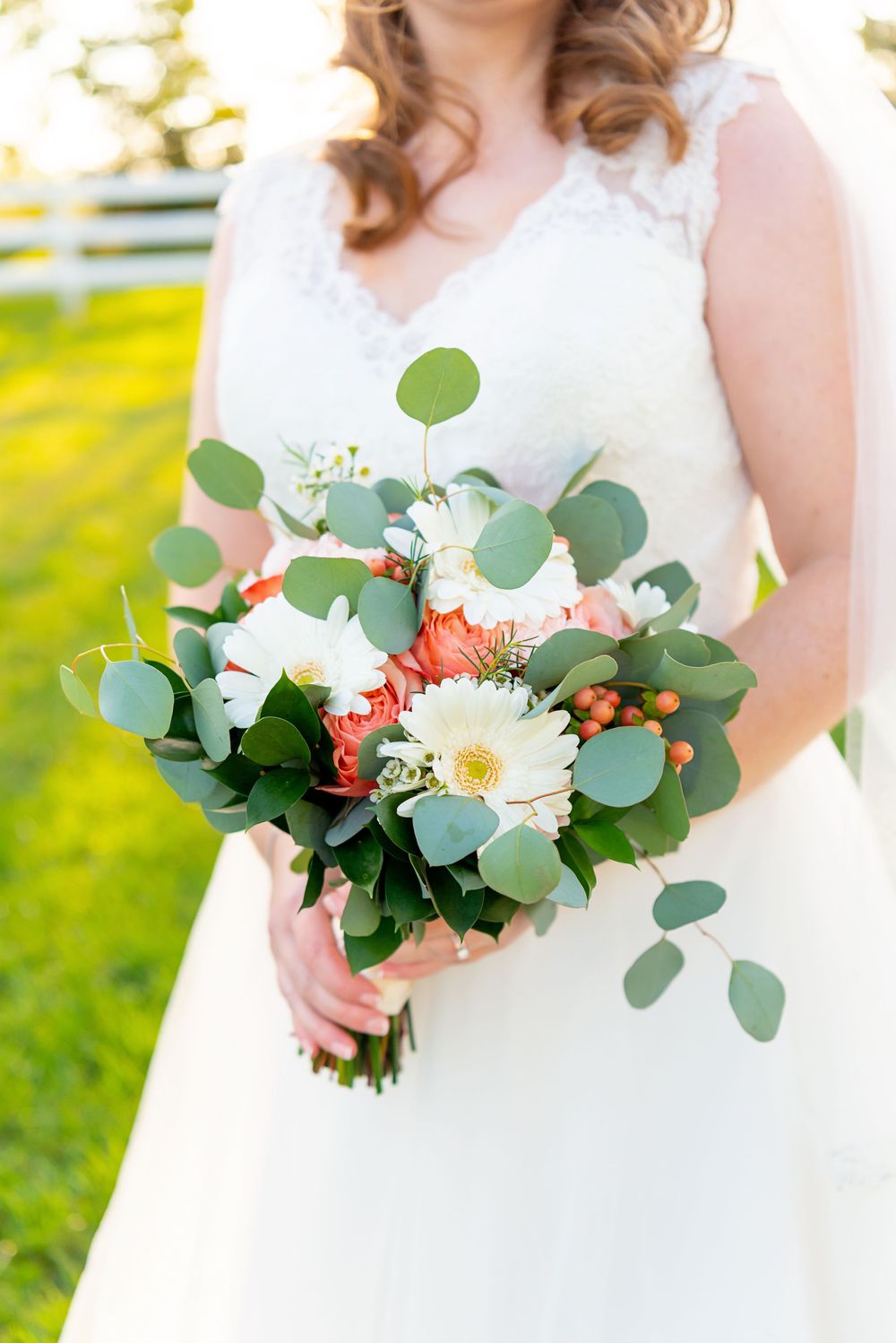 A white wedding bouquet features coral roses, white daisies, and eucalyptus greenery held against a white dress.