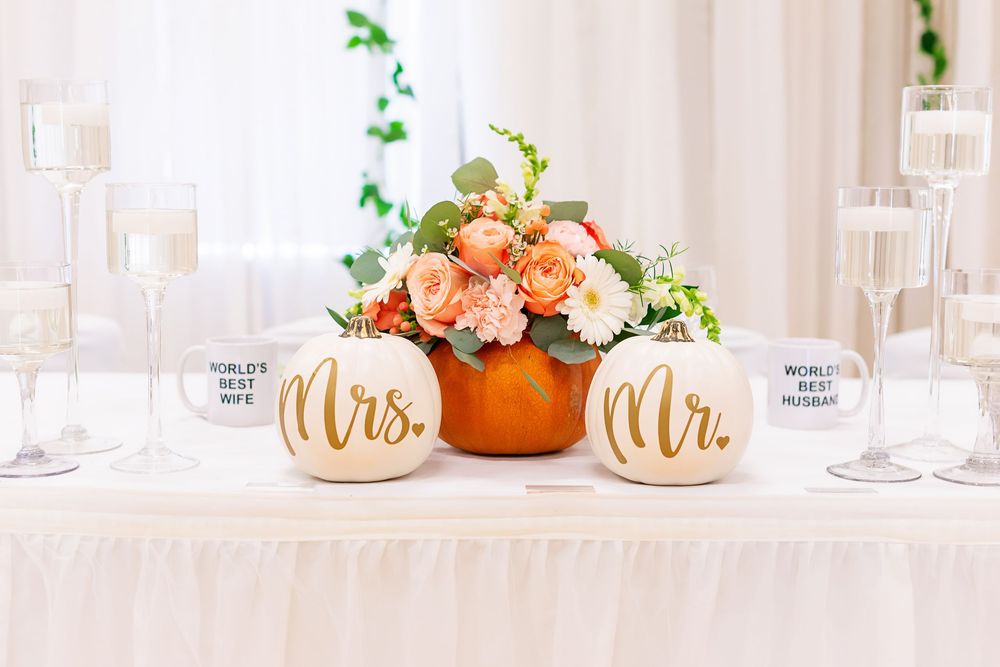 Wedding table decor with orange floral centerpiece and white pumpkins labeled Mr and Mrs beside champagne glasses.