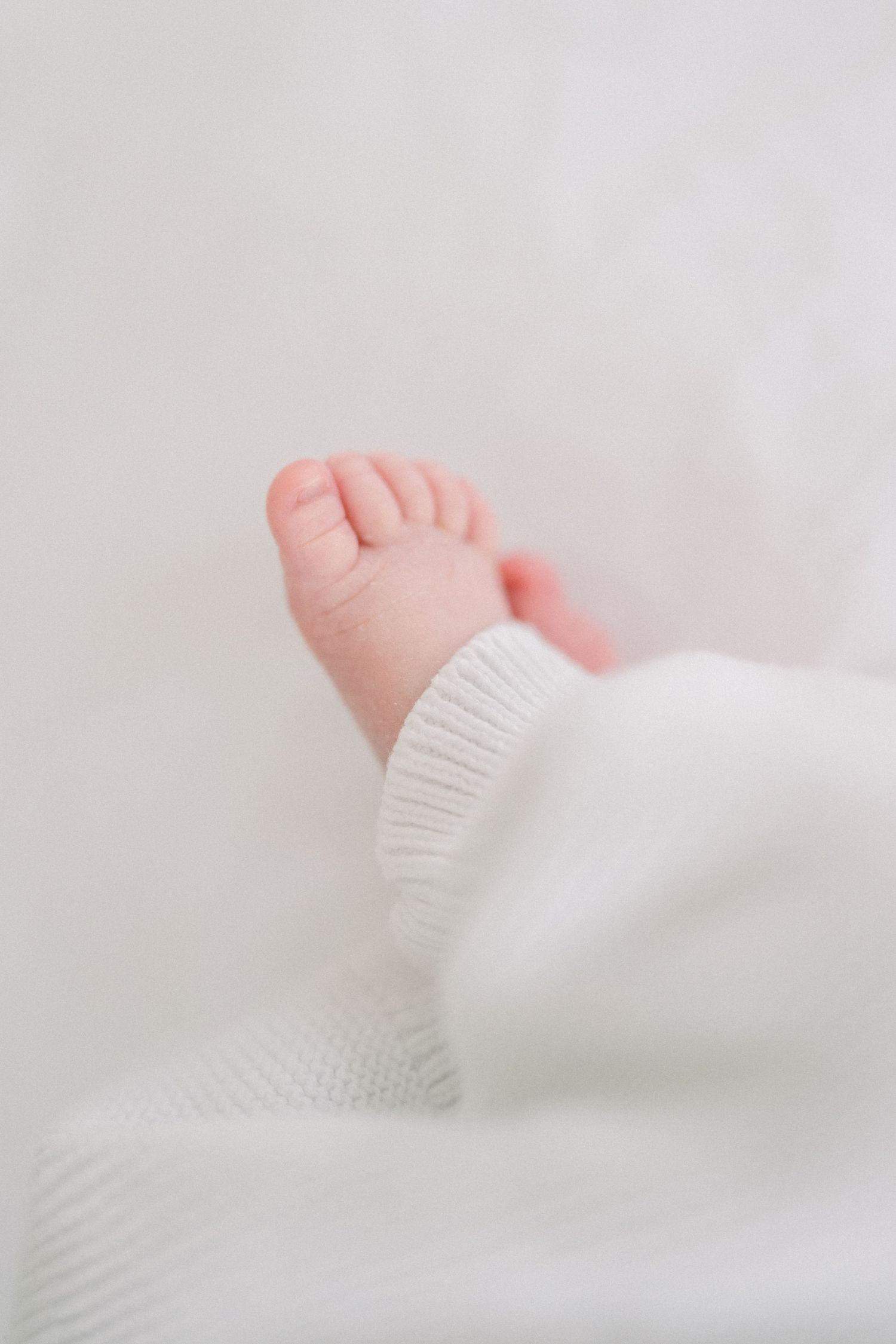 A tiny baby foot wearing a white knit sock rests on soft white bedding.