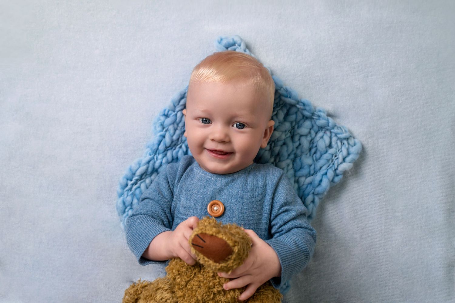 A baby in blue clothing plays and smiles while sitting with a teddy bear on a light gray background.