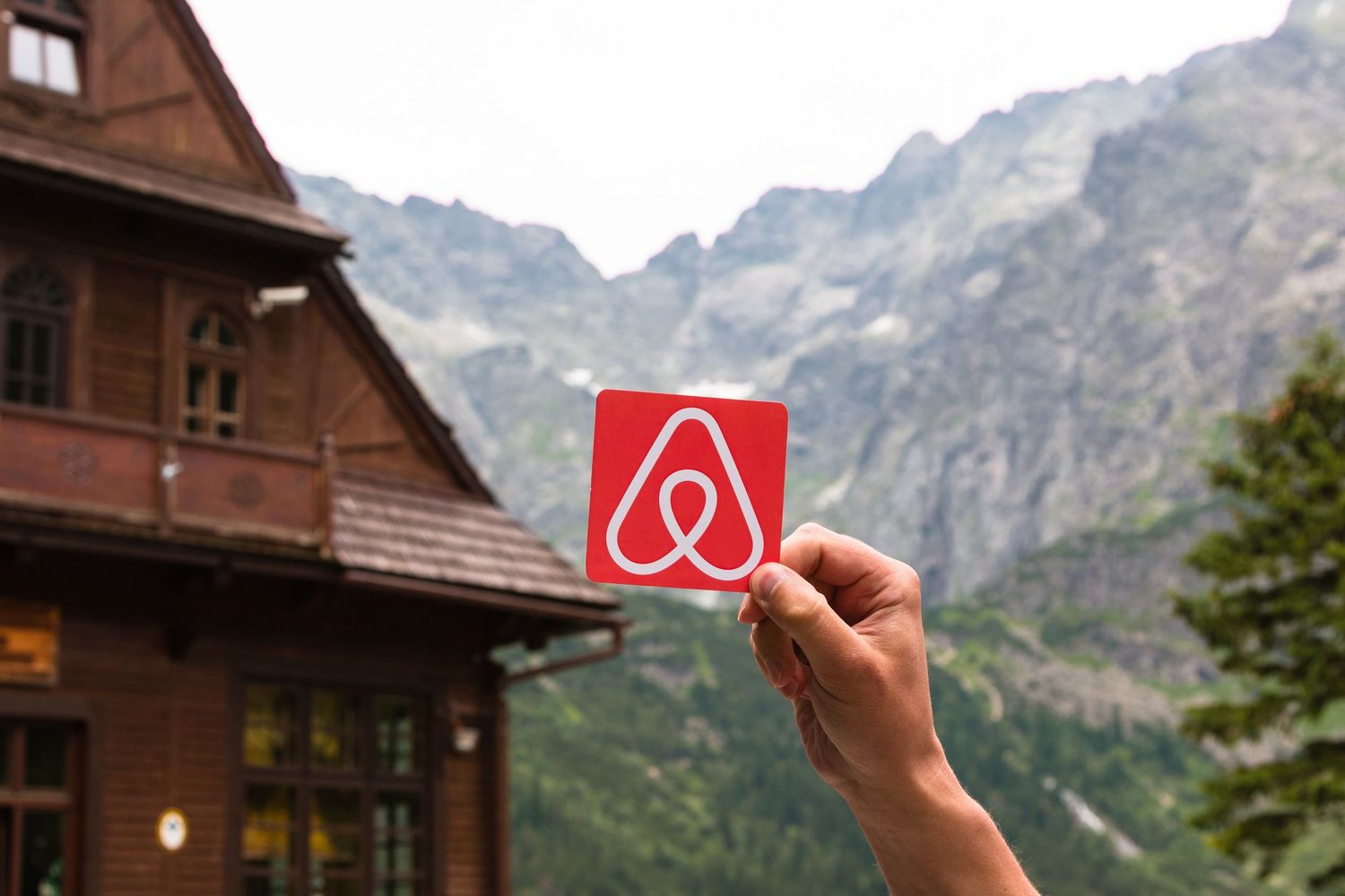 Hand holding up a red Airbnb logo card against a backdrop of rustic cabins and mountain peaks.