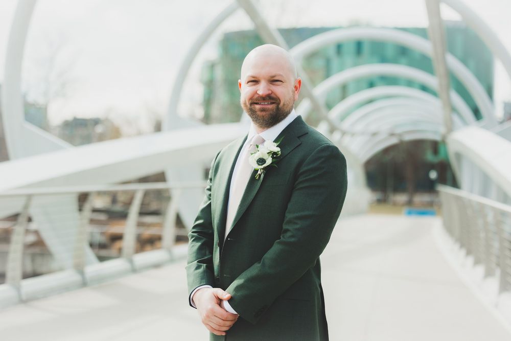 A groom in a dark suit stands on a riverside walkway bridge with curved green architectural elements in the background.