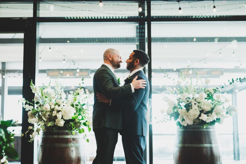 Two grooms in suits share an intimate wedding moment between floral arrangements in a bright, window-lit at the District Winery DC.