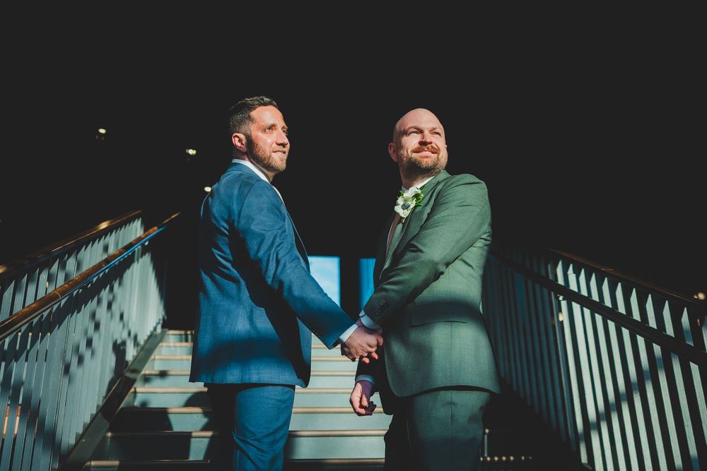 Two grooms in contrasting blue and green suits stand together on stairs while dramatic shadows create geometric patterns.