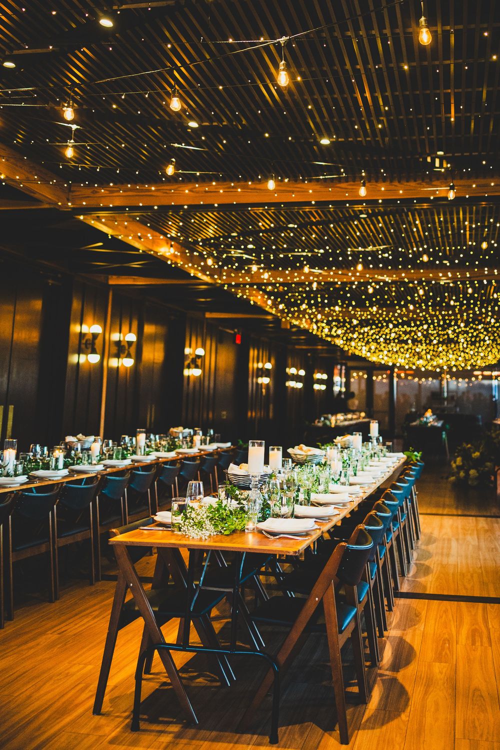 Modern restaurant interior with string lights overhead and long wooden tables with blue chairs creating an elegant ambiance.
