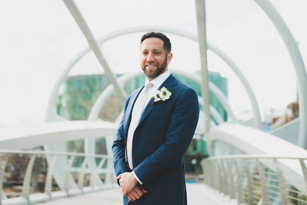 A groom in a navy suit stands on a modern white bridge walkway with architectural arches in the background.