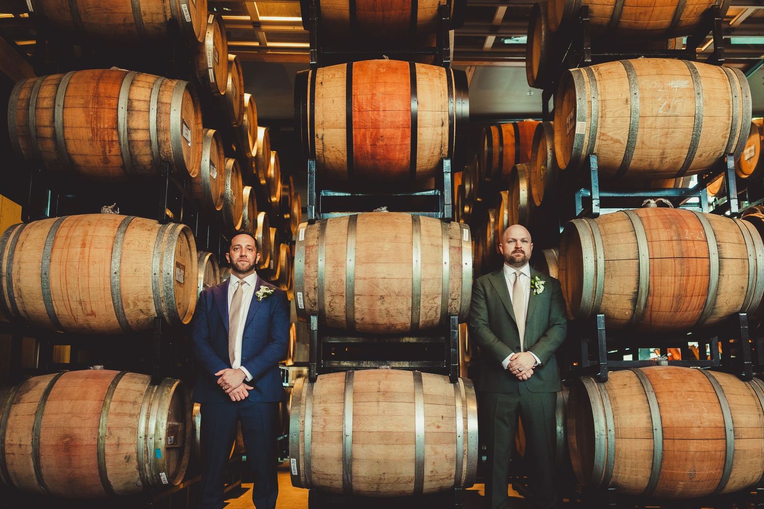 Two grooms stand in front of stacked wooden wine barrels in a dimly lit winery cellar.