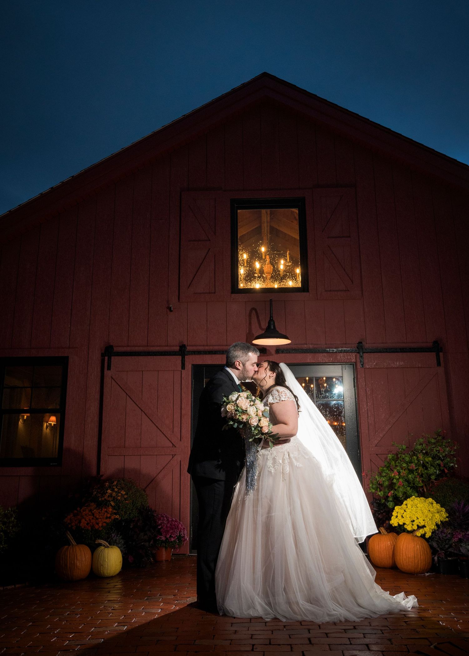 A romantic wedding couple poses in front of a rustic red barn at dusk with glowing windows and autumn pumpkins.