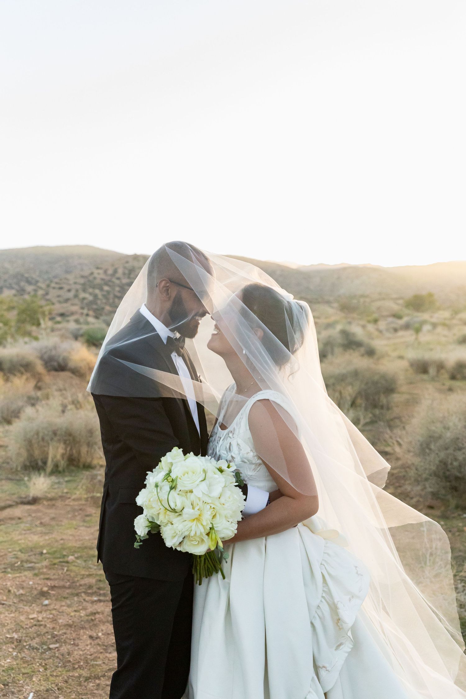 A couple shares a romantic moment at sunset in a desert landscape, with the bride holding a white bouquet.