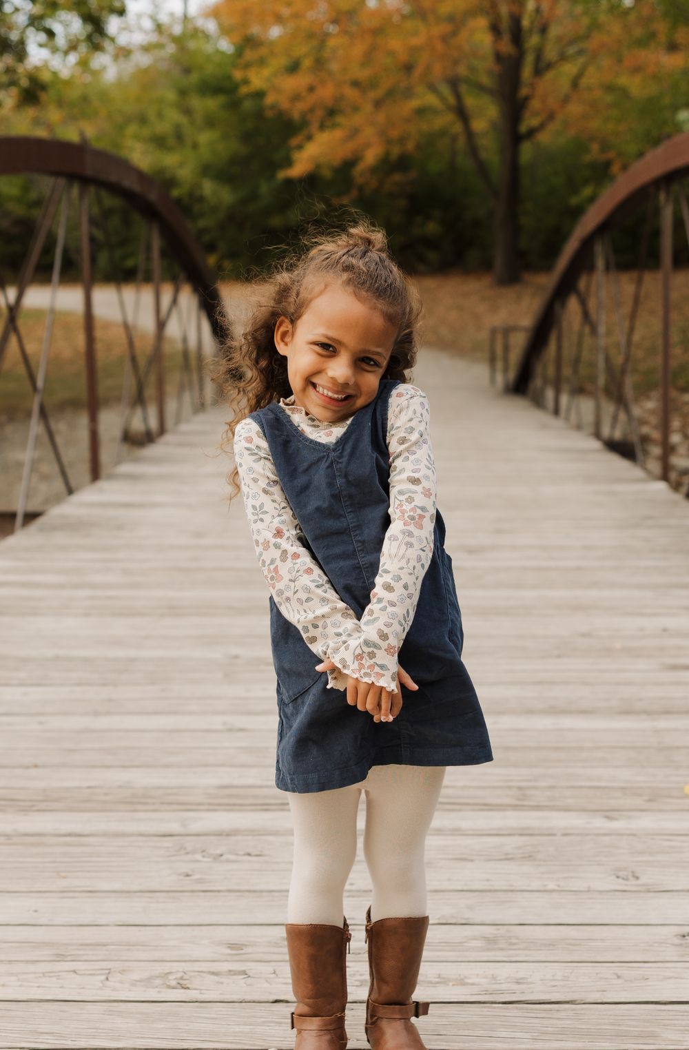 Child in navy vest and floral top poses playfully on wooden boardwalk during autumn season.