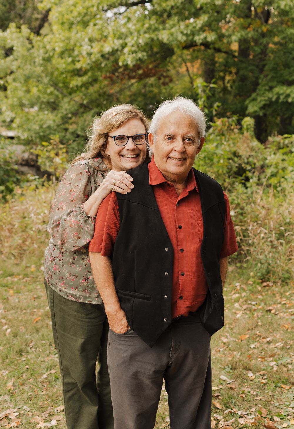 Two people wearing casual attire share warm embraces while posing together in a natural outdoor setting with trees.
