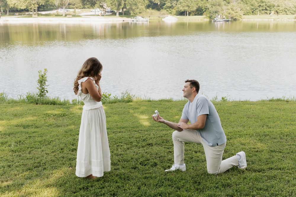 A romantic lakeside proposal scene unfolds on a grassy lawn during a sunny summer day.