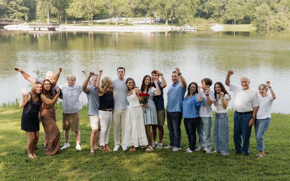 Large group of people standing together outdoors by a lake wearing casual attire for a group photo.