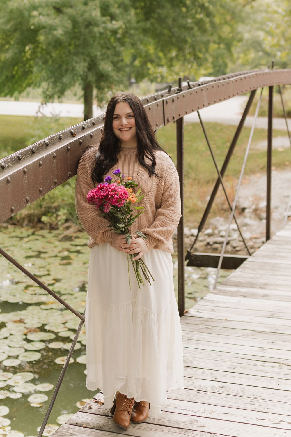 A person in a white dress and beige cardigan sits on a wooden dock holding pink flowers near a serene pond.