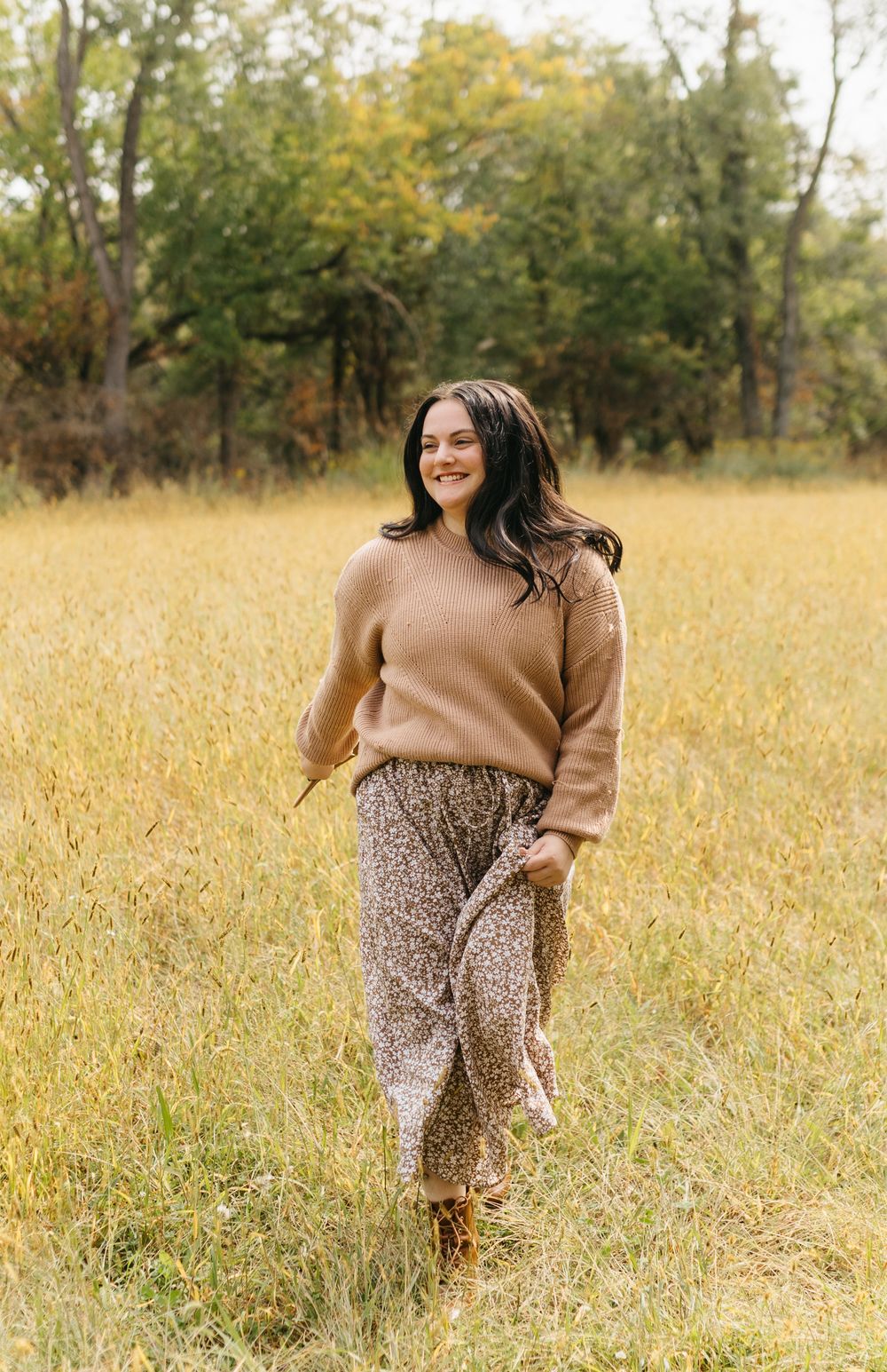 A woman in a brown sweater and leopard print skirt walks through a golden autumn field at sunset.