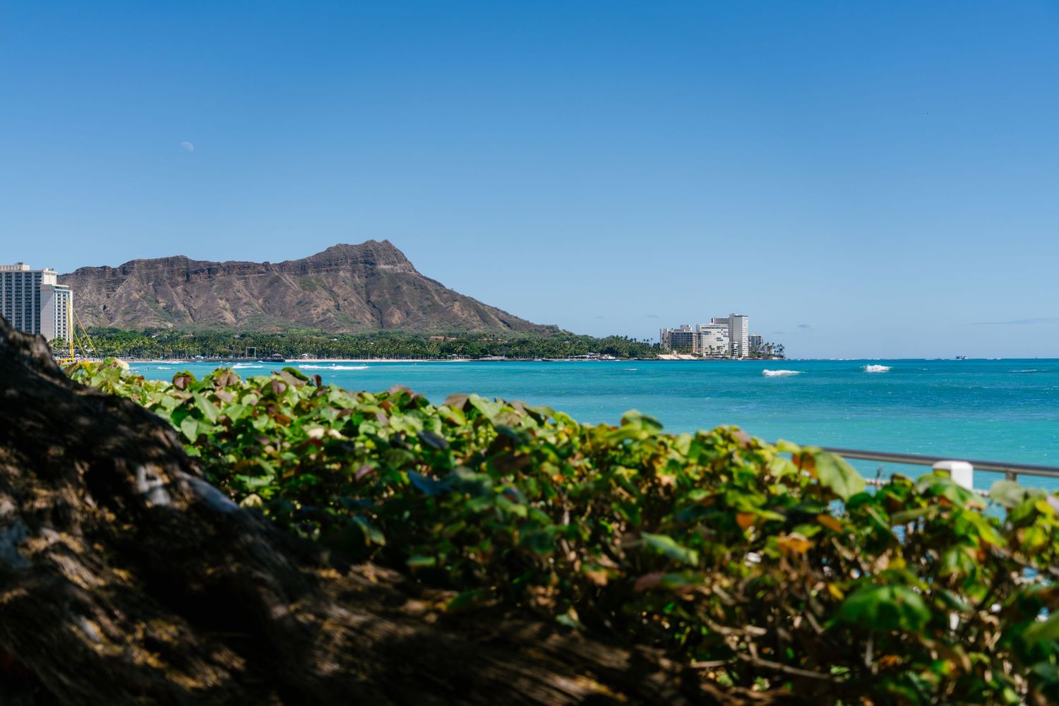 Turquoise waters of Waikiki Beach with Diamond Head crater and hotels in the background under a clear blue sky in front of Halekulani Hotel.