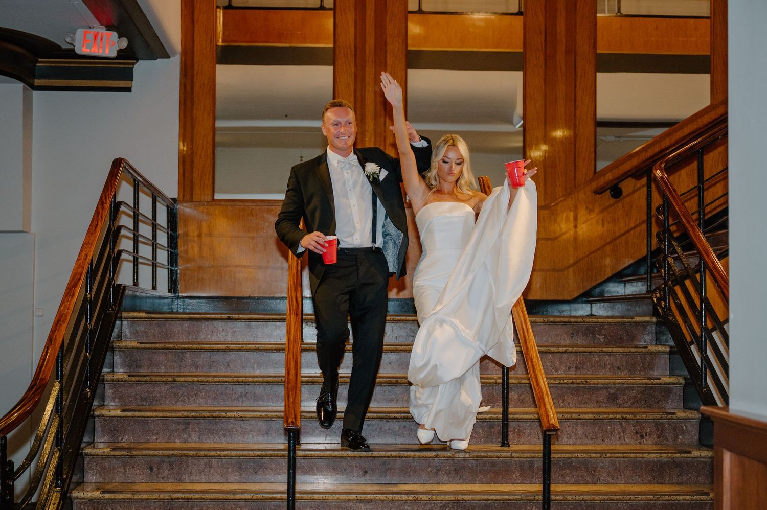 A wedding couple walks up an elegant wooden staircase in a historic building with warm lighting.