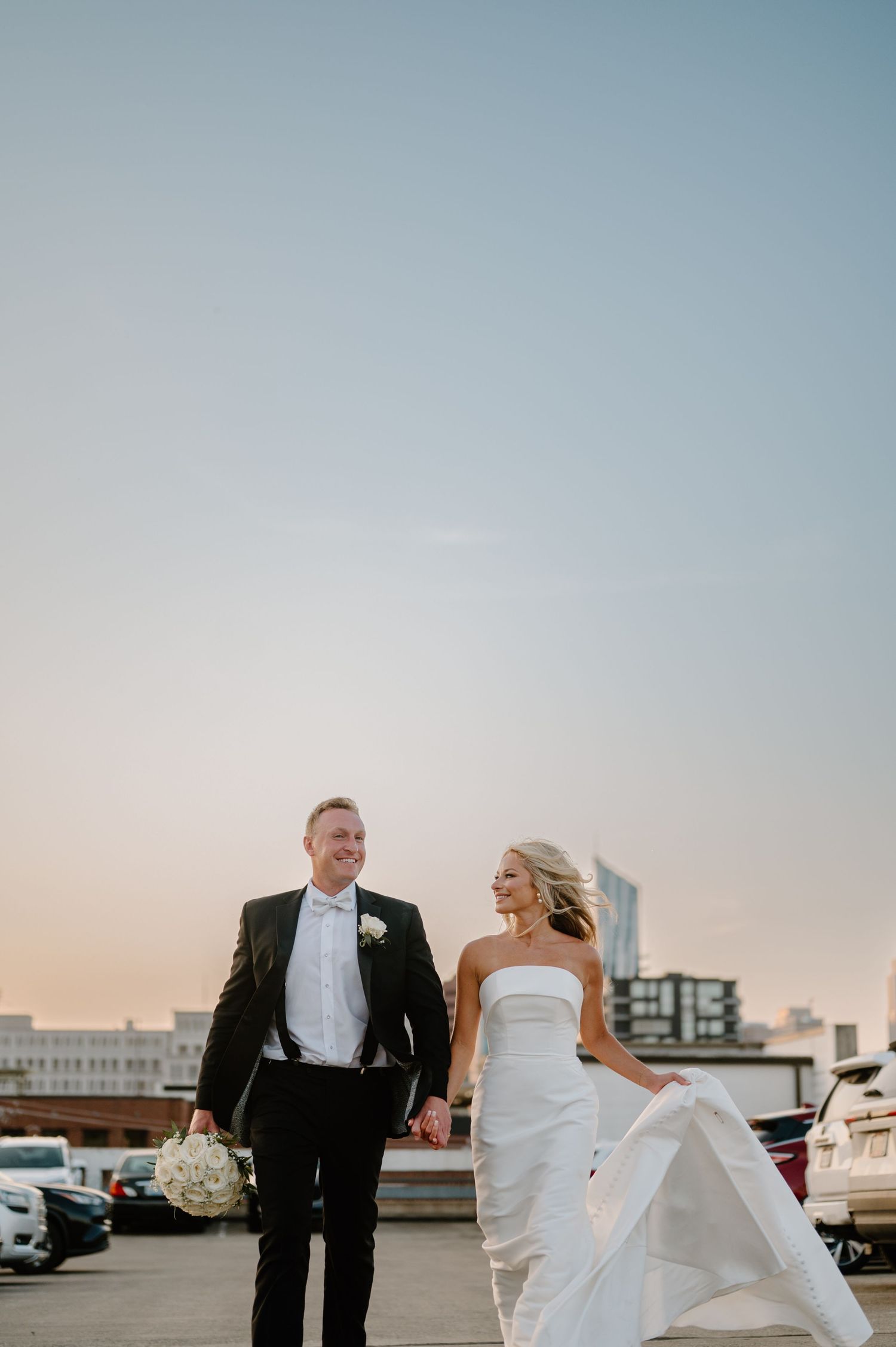 A couple in formal wedding attire walk and laugh together on a rooftop with a city skyline in the background.