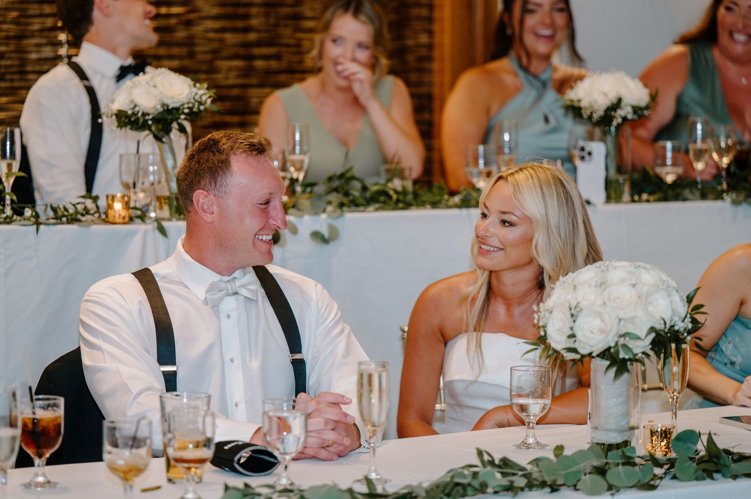 A wedding reception table decorated with white flowers and glassware as guests enjoy dinner and conversation.