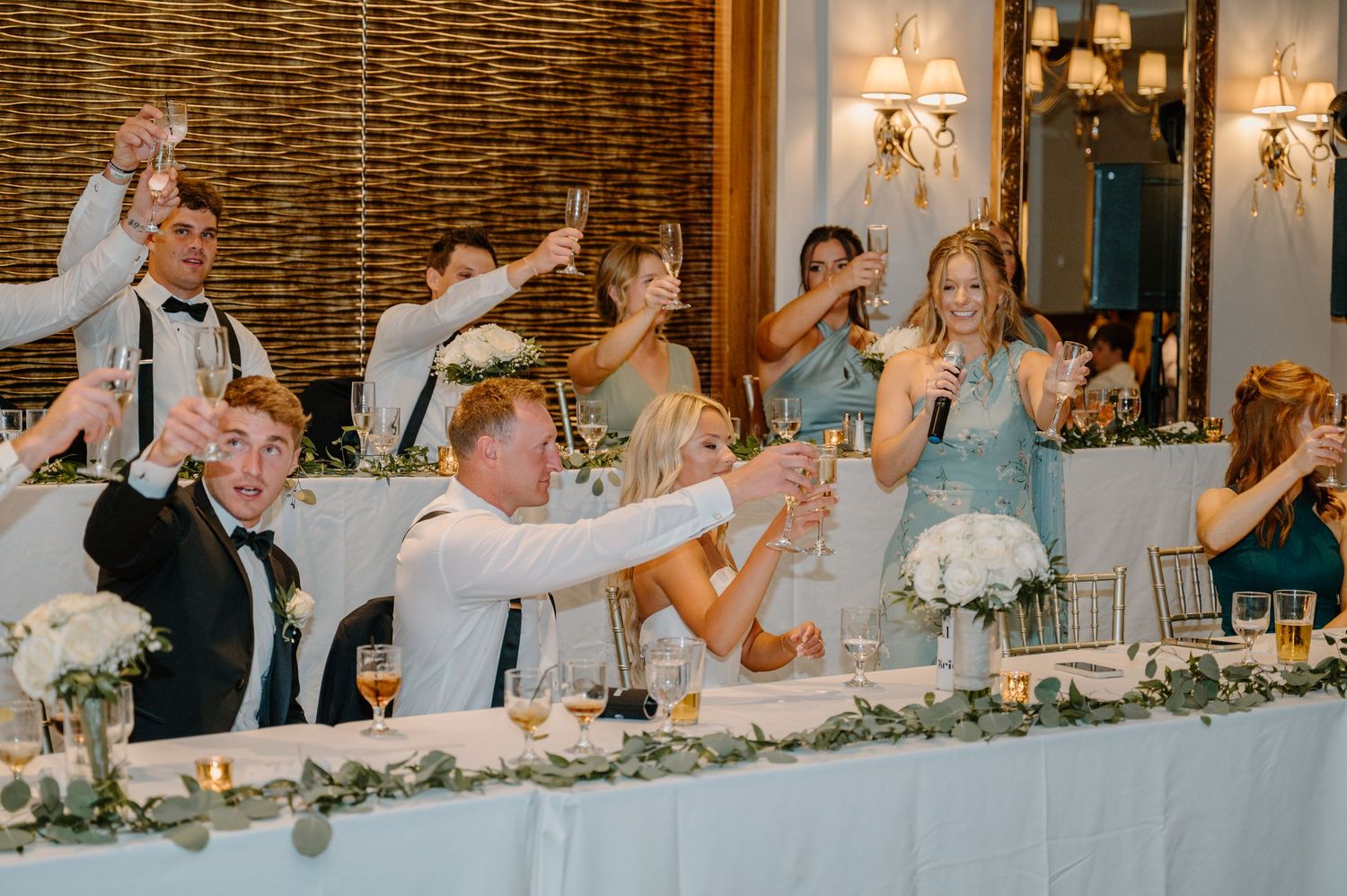 Wedding party guests raise their glasses in celebration at an elegant reception with brick walls and floral centerpieces.